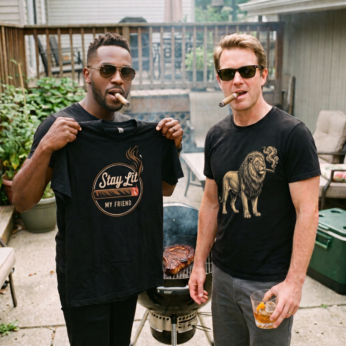 Two men standing outdoors in a backyard patio setting, both wearing black Legacy Layers Cigar t-shirts and smoking cigars. The man on the left holds up a black t-shirt that reads 'Stay-Fit MY FRIEND' with a cigar graphic.